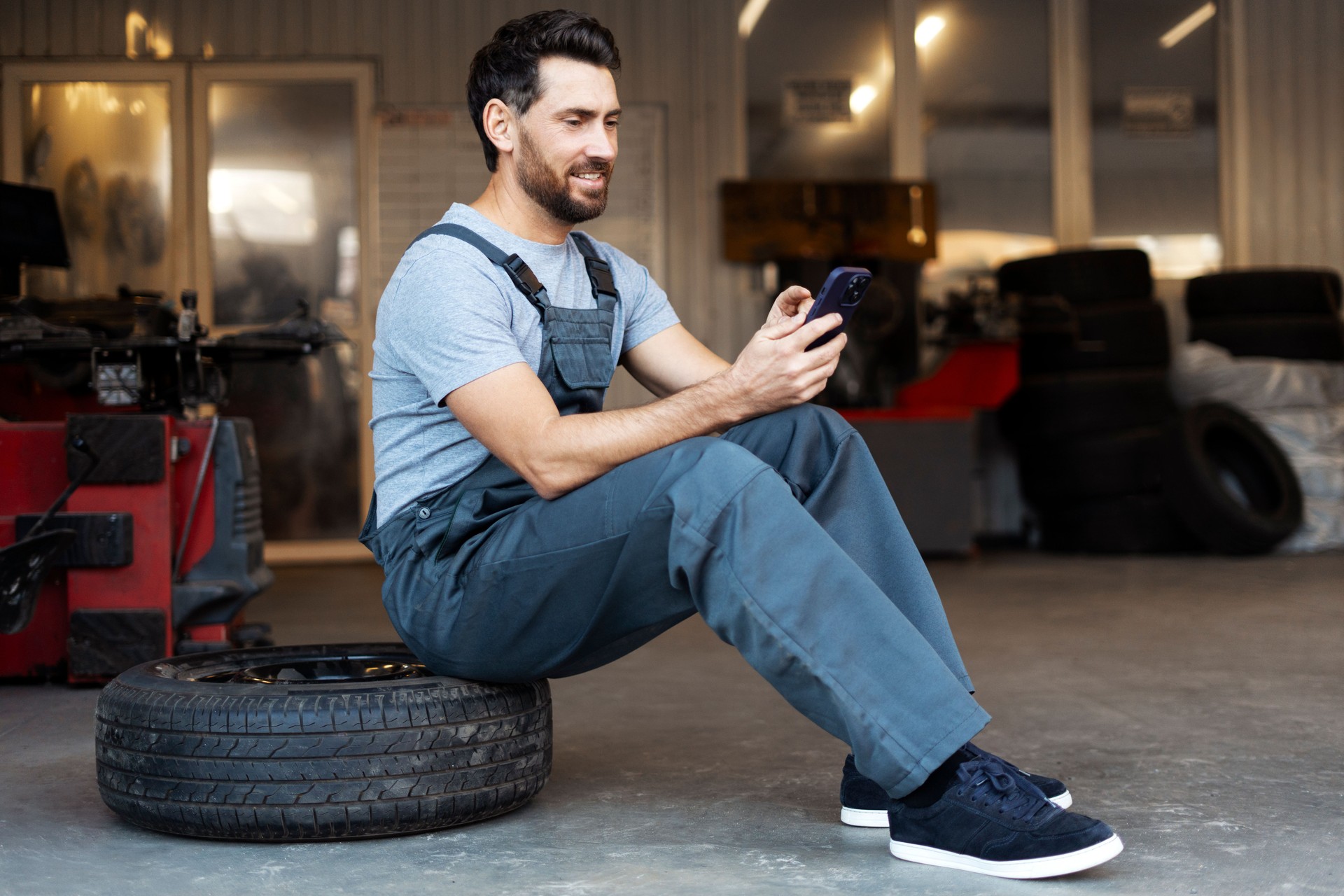 Mechanic sitting on tire browsing on mobile phone in auto repair shop